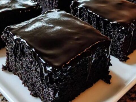 A close-up of a fork sinking into a moist dark chocolate sheet cake square with glossy glaze.