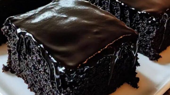 A close-up of a fork sinking into a moist dark chocolate sheet cake square with glossy glaze.