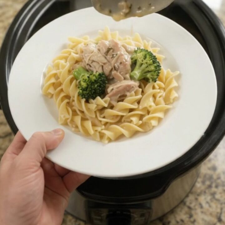 Creamy slow cooker chicken and broccoli in a black crockpot, with a spoon lifting a saucy bite.