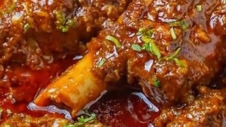 A close-up shot of tender meat shanks covered in a thick, glossy red curry sauce in a metal bowl.