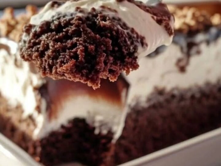 A close-up of a silver spoon scooping into a decadent chocolate cake topped with whipped cream and fudge.
