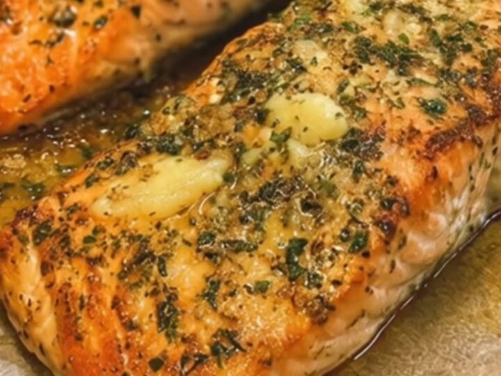 Two baked salmon fillets coated in melted garlic herb butter on parchment paper.