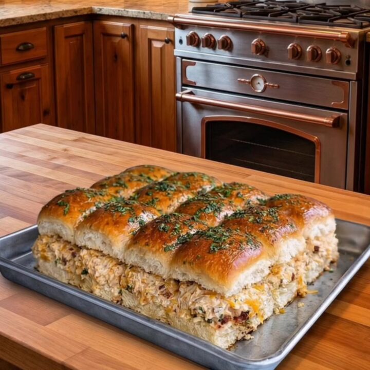A close-up of cheesy chicken bacon sliders on a metal pan in a rustic kitchen.