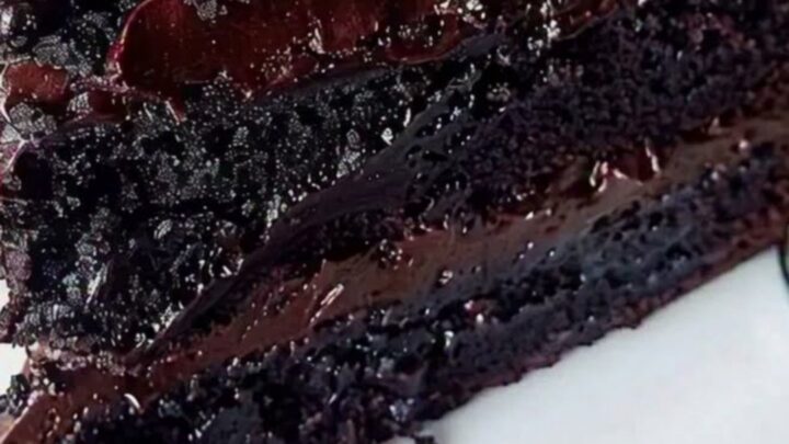 A close-up of a three-layer chocolate fudge cake slice with glossy ganache dripping down the sides on a white plate.