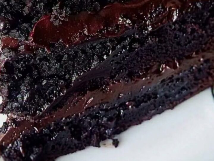 A close-up of a three-layer chocolate fudge cake slice with glossy ganache dripping down the sides on a white plate.