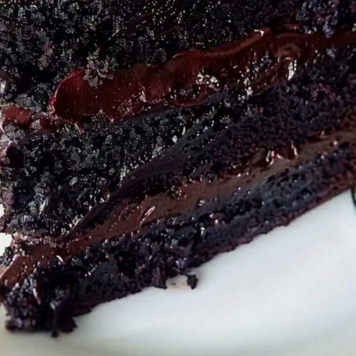 A close-up of a three-layer chocolate fudge cake slice with glossy ganache dripping down the sides on a white plate.