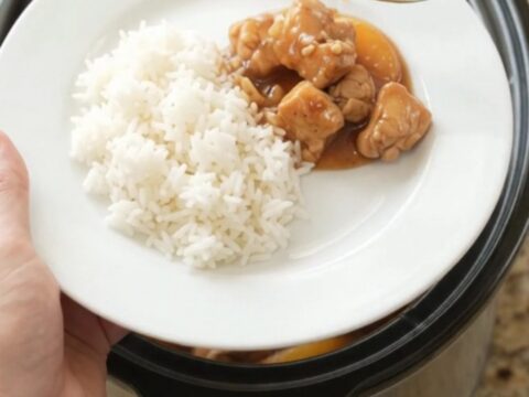 A slow cooker filled with peach teriyaki chicken bites, garnished with sesame seeds, on a granite countertop.
