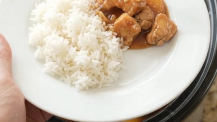 A slow cooker filled with peach teriyaki chicken bites, garnished with sesame seeds, on a granite countertop.