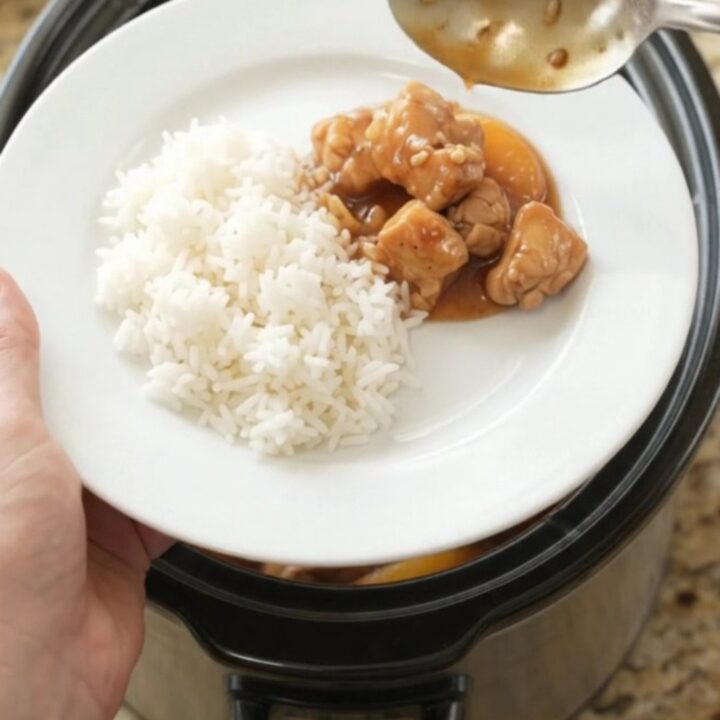 A slow cooker filled with peach teriyaki chicken bites, garnished with sesame seeds, on a granite countertop.