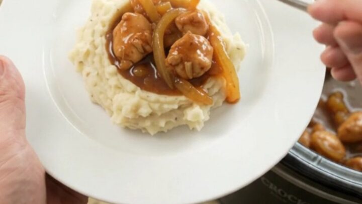A slow cooker filled with glazed chicken bites in sticky apricot bourbon sauce, topped with caramelized onion ribbons.