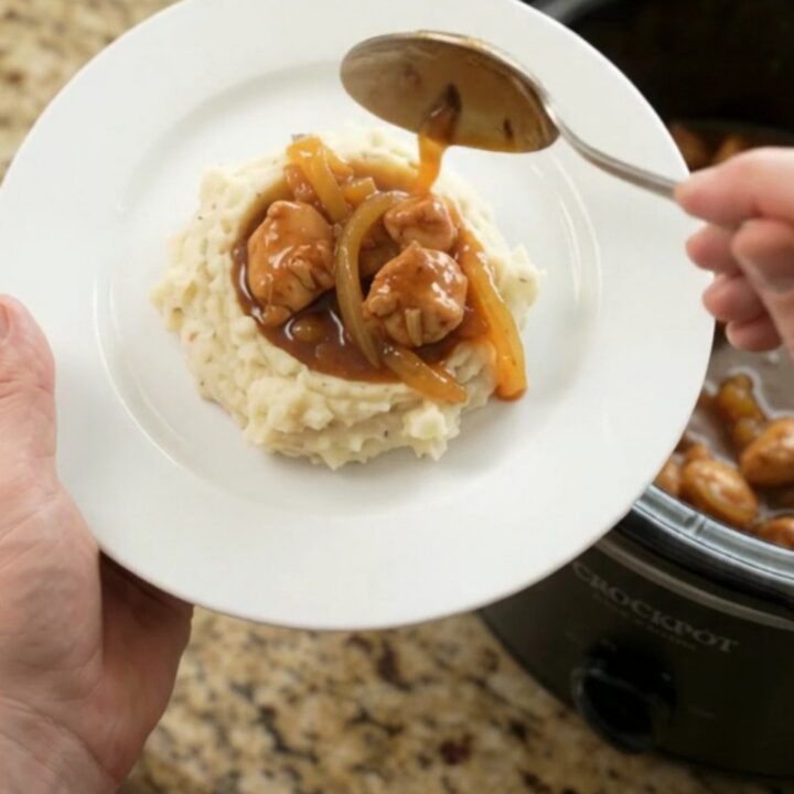 A slow cooker filled with glazed chicken bites in sticky apricot bourbon sauce, topped with caramelized onion ribbons.