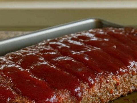 A glistening honey BBQ meatloaf served in a baking pan on a wire rack with a sunlit window in the background.