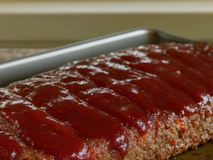 A glistening honey BBQ meatloaf served in a baking pan on a wire rack with a sunlit window in the background.