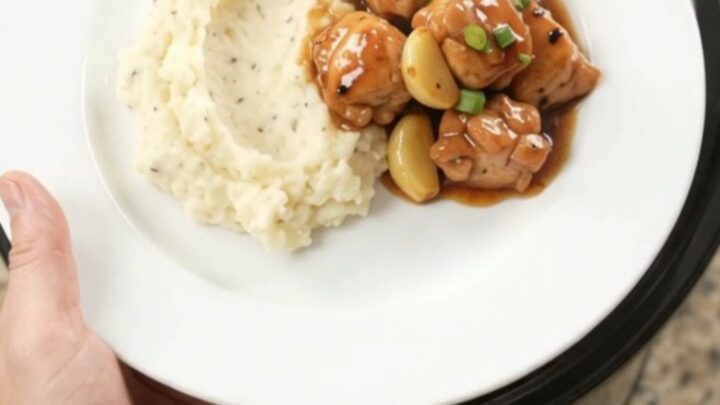 Slow cooker filled with glazed chicken bites, garlic cloves, and scallions in a dark bourbon sauce.