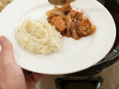 Slow cooker full of glazed chicken bites and caramelized onions with a spoon lifting a portion