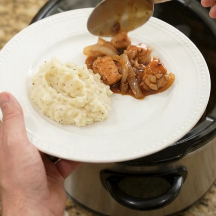 Slow cooker full of glazed chicken bites and caramelized onions with a spoon lifting a portion