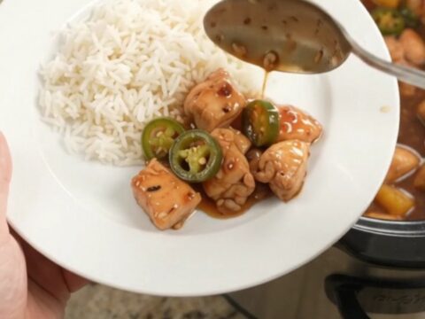 A slow cooker filled with glazed honey soy chicken bites, pineapple, and jalapeños, with steam rising.