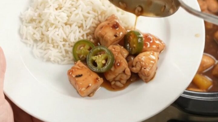 A slow cooker filled with glazed honey soy chicken bites, pineapple, and jalapeños, with steam rising.