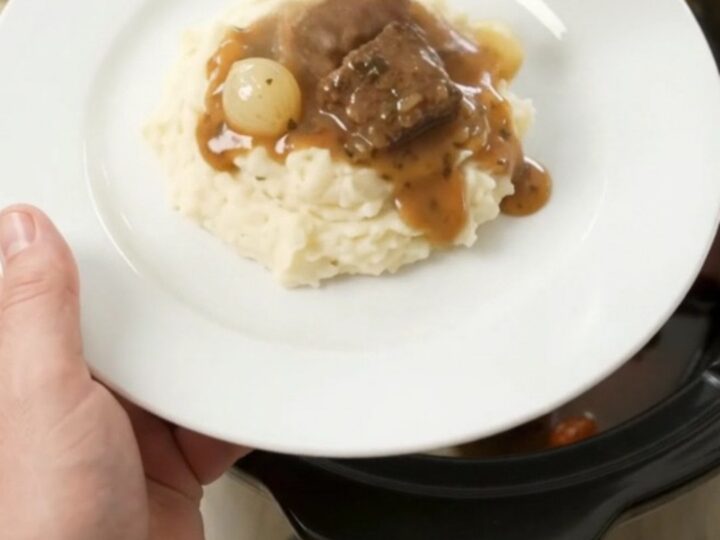 Slow cooker beef bites with pearl onions and carrots in a glossy butter au jus, topped with a spoonful of sauce.
