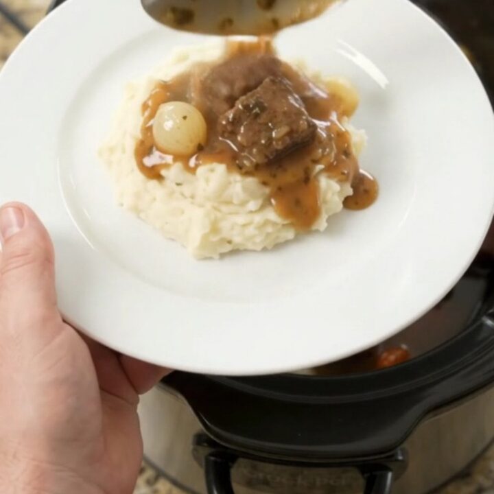 Slow cooker beef bites with pearl onions and carrots in a glossy butter au jus, topped with a spoonful of sauce.