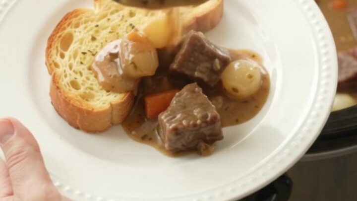 A slow cooker filled with tender beef chunks, pearl onions, and carrots in a creamy Gruyère sauce