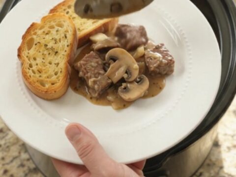 A slow cooker filled with creamy ribeye bites and mushrooms, topped with melted Gruyère