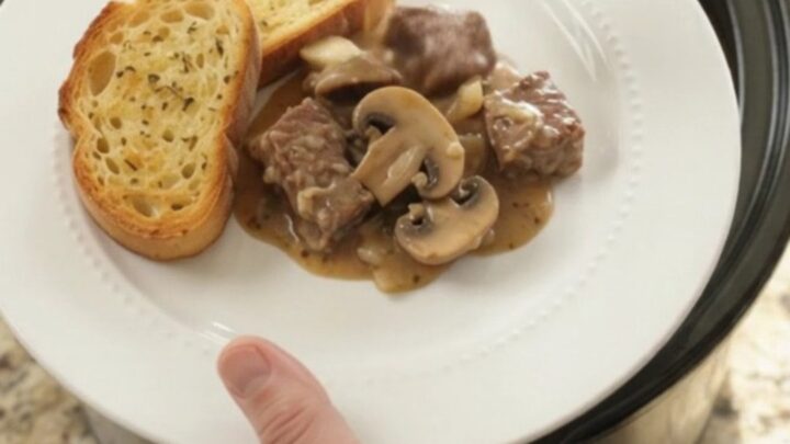 A slow cooker filled with creamy ribeye bites and mushrooms, topped with melted Gruyère