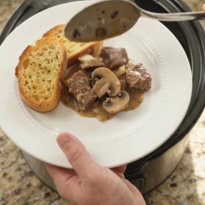 A slow cooker filled with creamy ribeye bites and mushrooms, topped with melted Gruyère