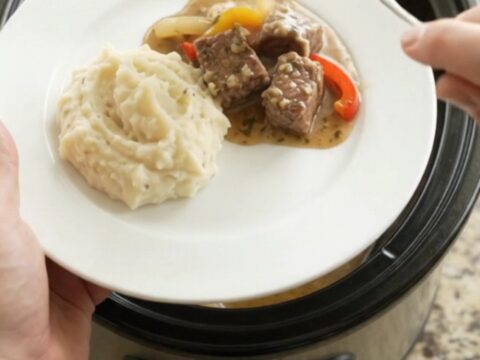 Slow-cooked ribeye bites with bell peppers and onions in a rich butter au jus in a black ceramic slow cooker.