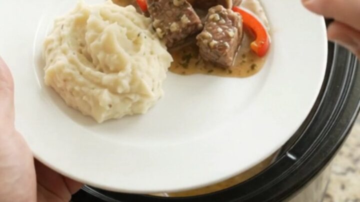 Slow-cooked ribeye bites with bell peppers and onions in a rich butter au jus in a black ceramic slow cooker.