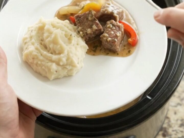 Slow-cooked ribeye bites with bell peppers and onions in a rich butter au jus in a black ceramic slow cooker.