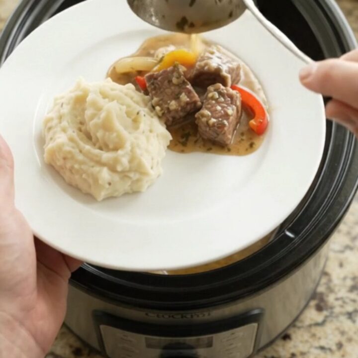 Slow-cooked ribeye bites with bell peppers and onions in a rich butter au jus in a black ceramic slow cooker.