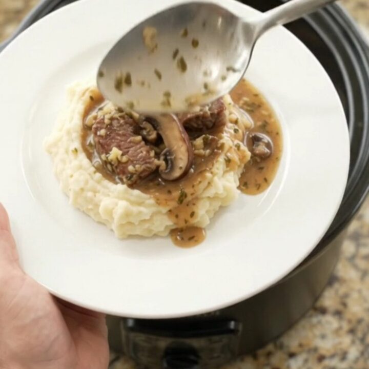 Slow cooker ribeye bites and portobello mushrooms in a rich garlic-herb butter au jus, served in a black crockpot.