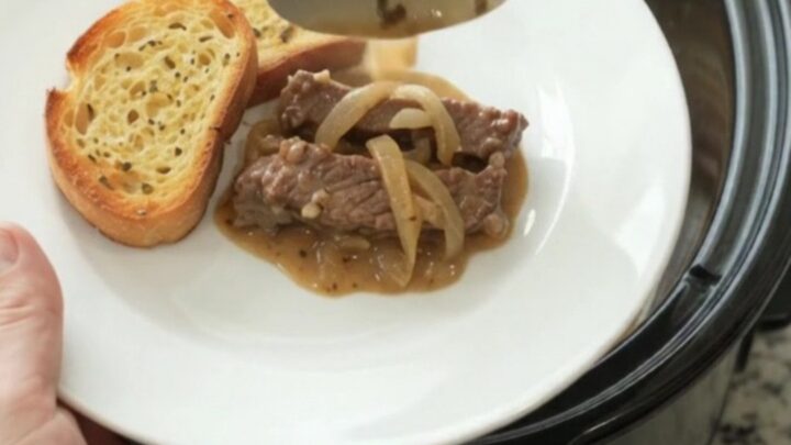 Slow cooker filled with tender flank steak and onions in a creamy Gruyère sauce, with steam rising