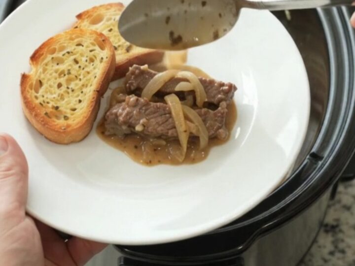 Slow cooker filled with tender flank steak and onions in a creamy Gruyère sauce, with steam rising