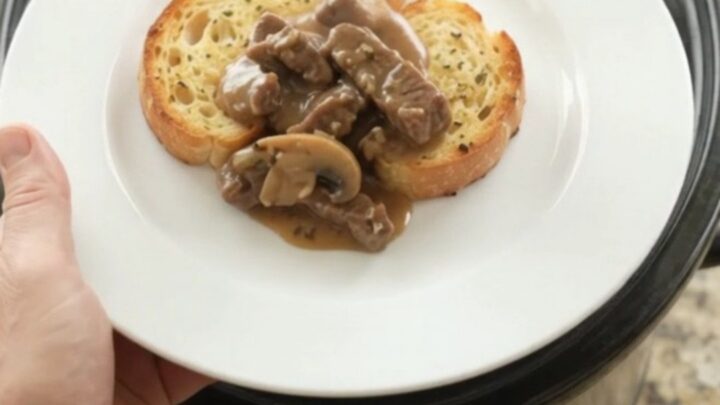 Slow cooker flank steak with mushrooms and shallots in a creamy Gruyère sauce, served on a white plate with brioche.