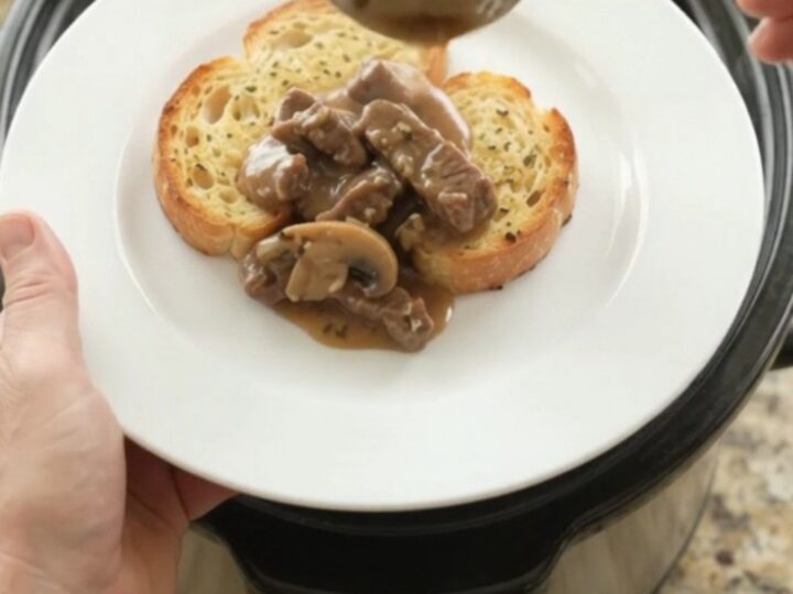 Slow cooker flank steak with mushrooms and shallots in a creamy Gruyère sauce, served on a white plate with brioche.