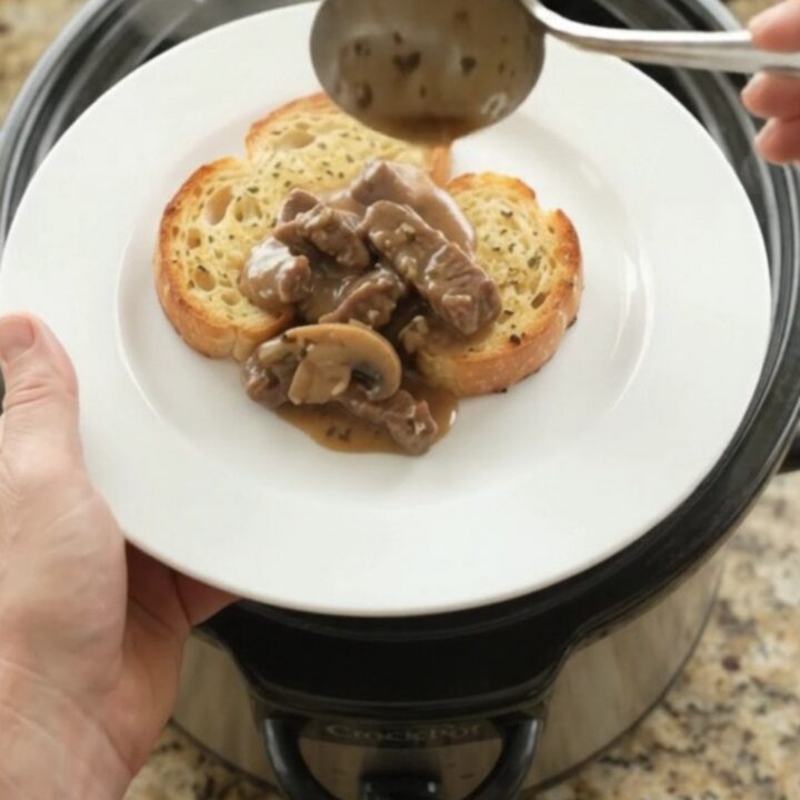 Slow cooker flank steak with mushrooms and shallots in a creamy Gruyère sauce, served on a white plate with brioche.