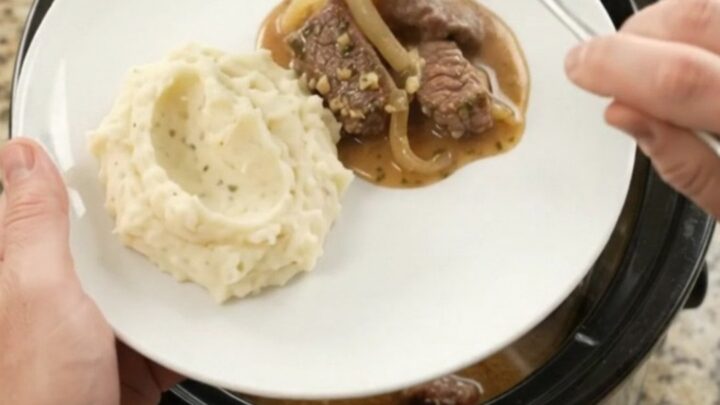 Slow cooker full of tender beef tips and caramelized onions in a glossy au jus, with steam rising