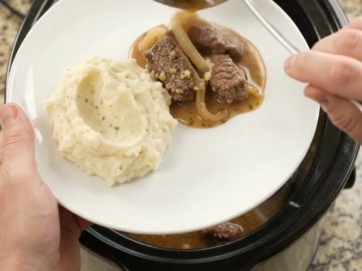 Slow cooker full of tender beef tips and caramelized onions in a glossy au jus, with steam rising