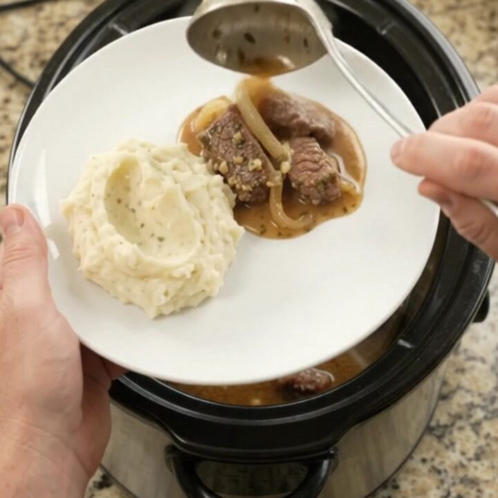 Slow cooker full of tender beef tips and caramelized onions in a glossy au jus, with steam rising