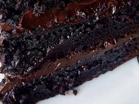 A triple-layer chocolate fudge cake on a white plate with chocolate chips nearby.
