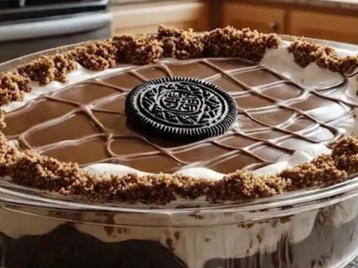 Chilled chocolate cake in a square glass dish topped with glossy ganache, piped grid, and crushed cookies.