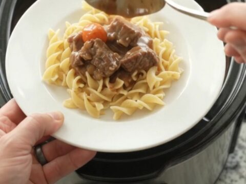 A slow cooker filled with tender beef tips and pearl onions in a rich brown gravy, with a spoon lifting a scoop.