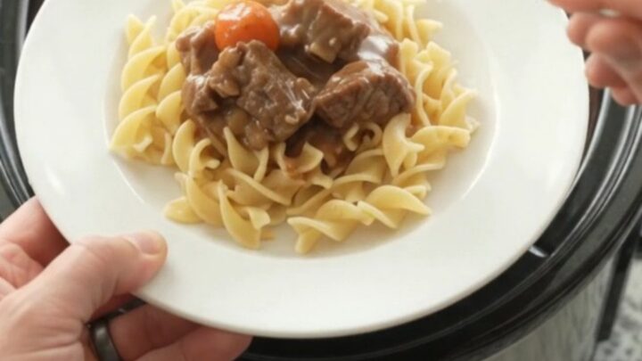 A slow cooker filled with tender beef tips and pearl onions in a rich brown gravy, with a spoon lifting a scoop.