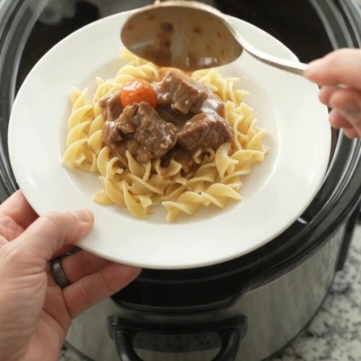 A slow cooker filled with tender beef tips and pearl onions in a rich brown gravy, with a spoon lifting a scoop.