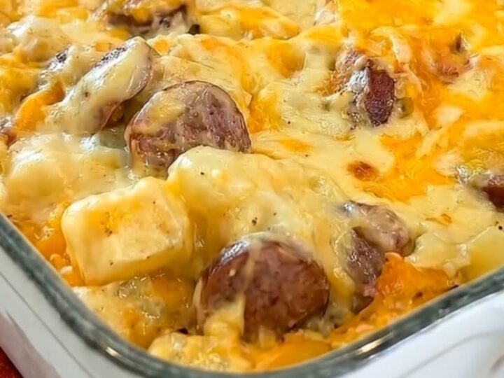 A golden, bubbly baked potato and sausage casserole in a white rectangular baking dish resting on a red cloth.