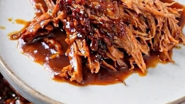 A rustic plate piled high with glistening, tender shredded beef dripping in a dark, rich barbecue sauce.