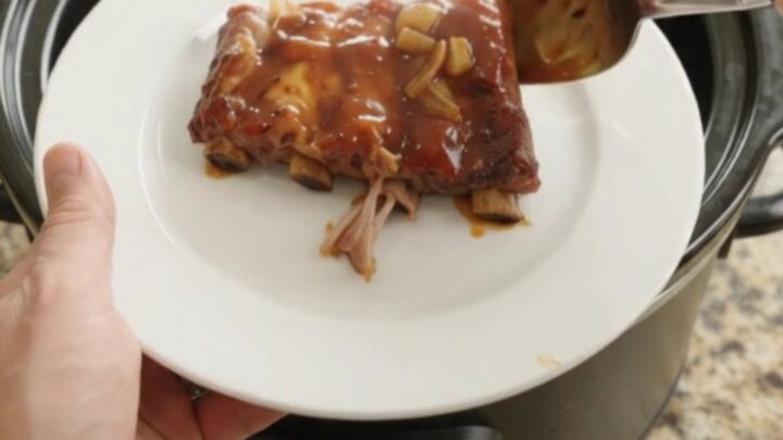 Root Beer BBQ Spare Ribs in slow cooker, glistening with sticky sauce and topped with caramelized onions.
