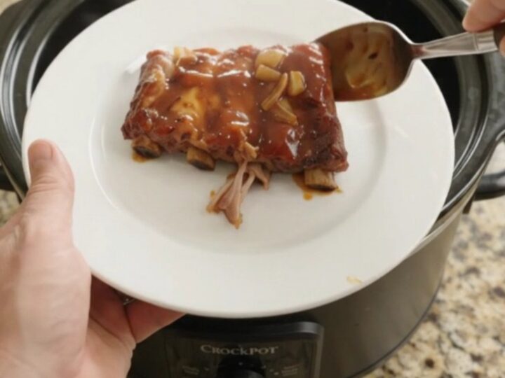 Root Beer BBQ Spare Ribs in slow cooker, glistening with sticky sauce and topped with caramelized onions.